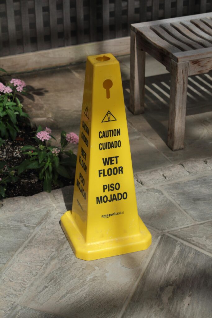 Yellow wet floor caution cone placed outdoors near a wooden bench and pink flowers.