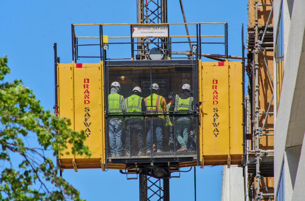 Group of construction workers in a lift at a building site, wearing safety vests and helmets.
