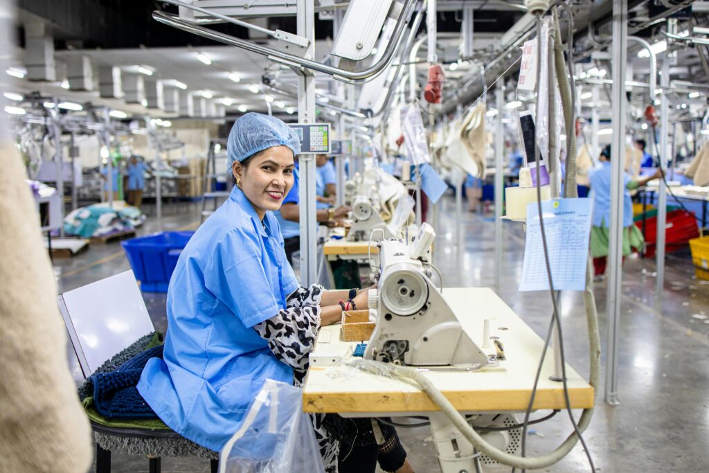 A female factory worker operating a sewing machine in a busy textile factory showing teamwork and industry.