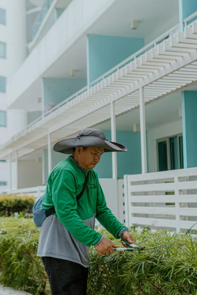 A gardener in a wide-brim hat trims hedges outside a contemporary building.